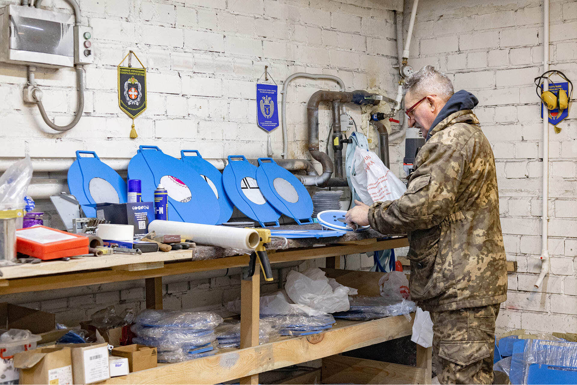 Рабочий в камуфляжной одежде собирает детали вакуумных камер на верстаке в производственном цехе, на столе — синие металлические крышки и инструменты.