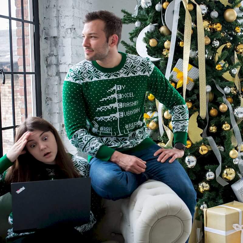 Фотография, изображающая мужчину и женщину, сидящих на светлом диване на фоне окна и новогодней елки. Мужчина в зелёном вязаном свитере с белыми узорами и надписью 'MY EXCEL CRASHES EVERYDAY #REF!' смотрит вправо, а женщина с темными волосами в похожем свитере, с удивленным выражением лица, держит ноутбук на коленях.