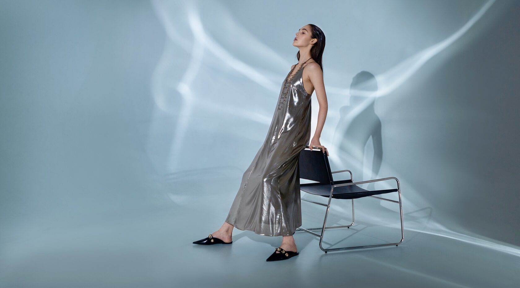 модель в длинном серебристом платье позирует, опираясь на стул на фоне циклорамы с голубым свечением