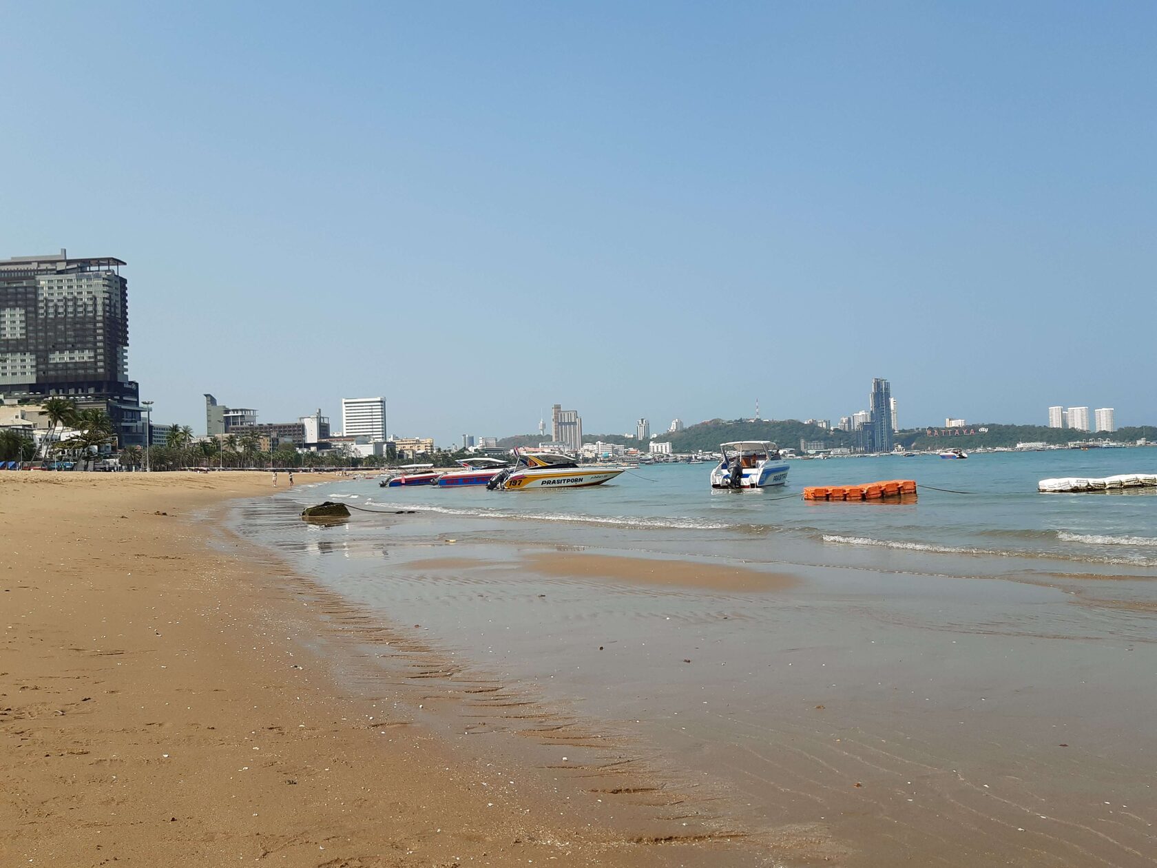 Песчаный пляж с лазурным морем, прогулочными катерами и панорамой современного курортного города. Спокойная береговая линия, чистый пляж и живописный морской пейзаж для отдыха, прогулок и пляжного туризма.