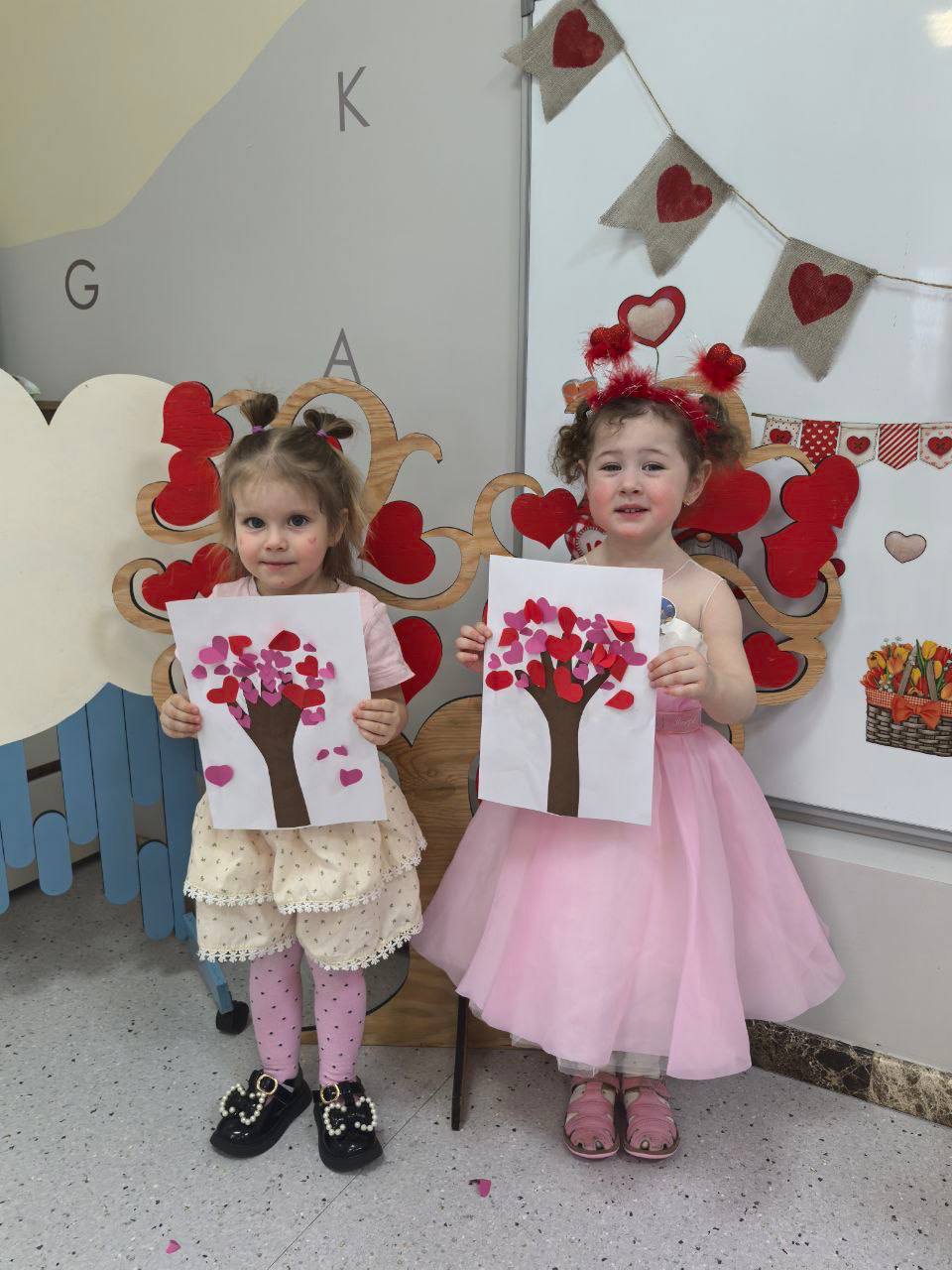 Две девочки в детском саду держат поделки с деревом из сердечек ко Дню святого Валентина
