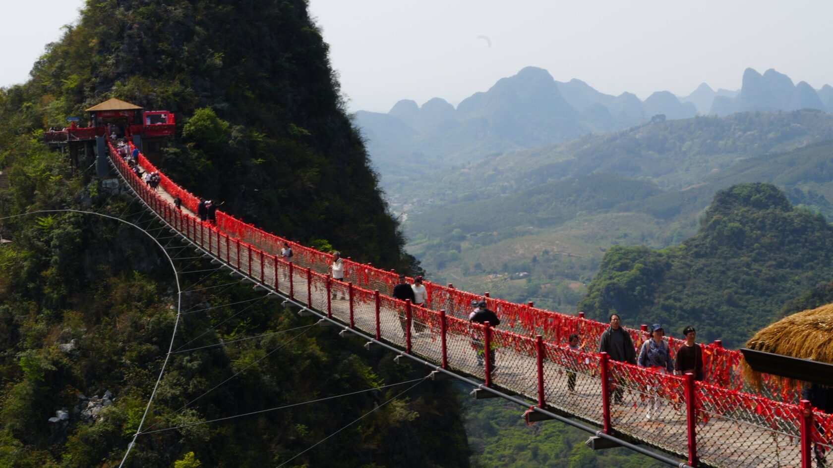 Подвесной мост с красными ограждениями на пике Жуи: туристы идут над ущельем, панорама карстовых гор Китая