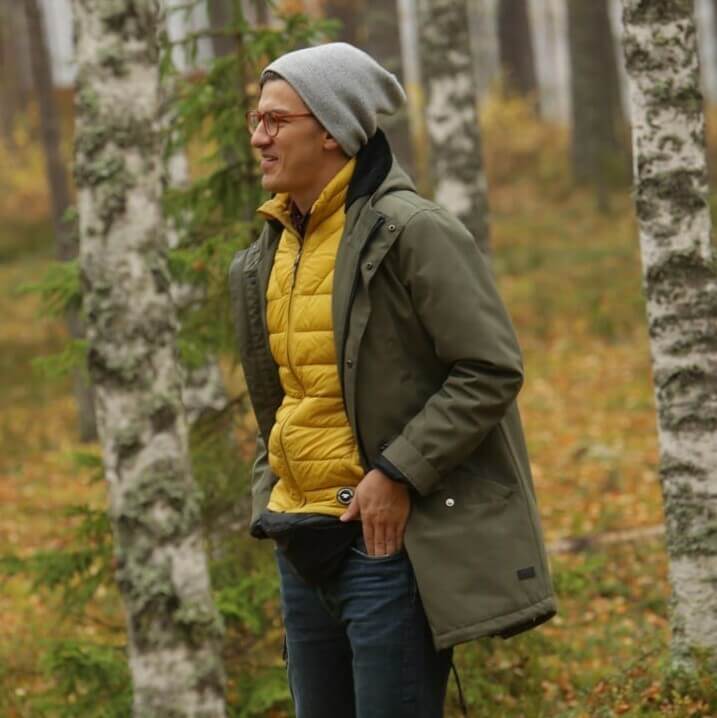 Фотография улыбающегося мужчины в очках с прозрачной оправой и серой шапке, в зелёной куртке и жёлтой жилетке, стоящего в осеннем лесу с размытыми деревьями на заднем плане. Мужчина смотрит влево и держит руки в карманах.