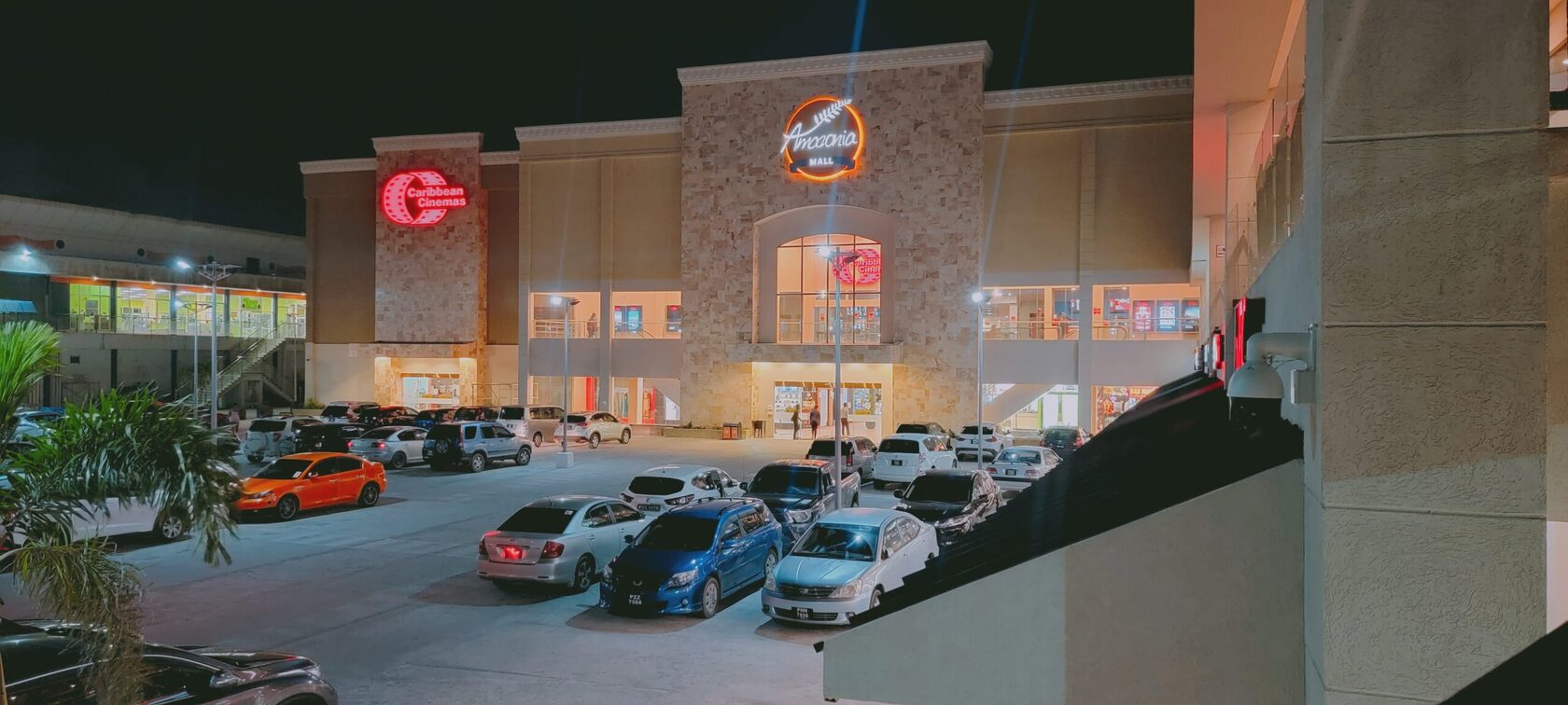 A wide-angle, nighttime shot of the exterior of a shopping mall. The main entrance is labeled with a neon sign that says Amazonia Mall. A red neon sign for Caribbean Cinemas is visible on the left. The parking lot in front is filled with cars, and the bui