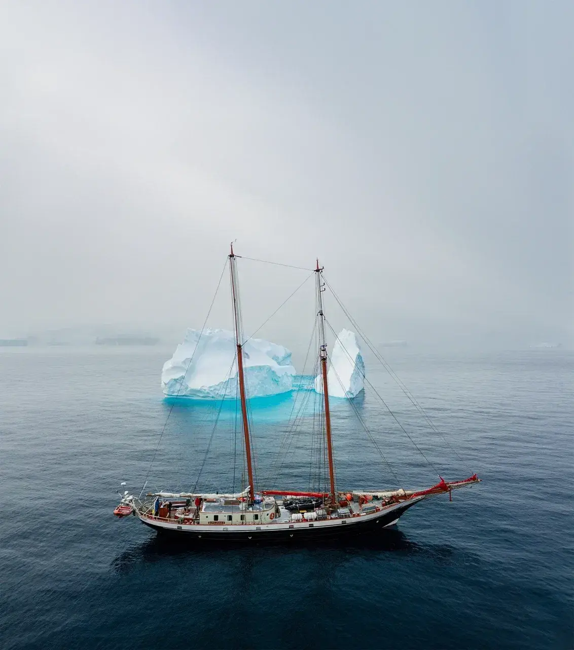 Sailing schooner navigating Antarctic waters near a massive iceberg during a luxury polar expedition.