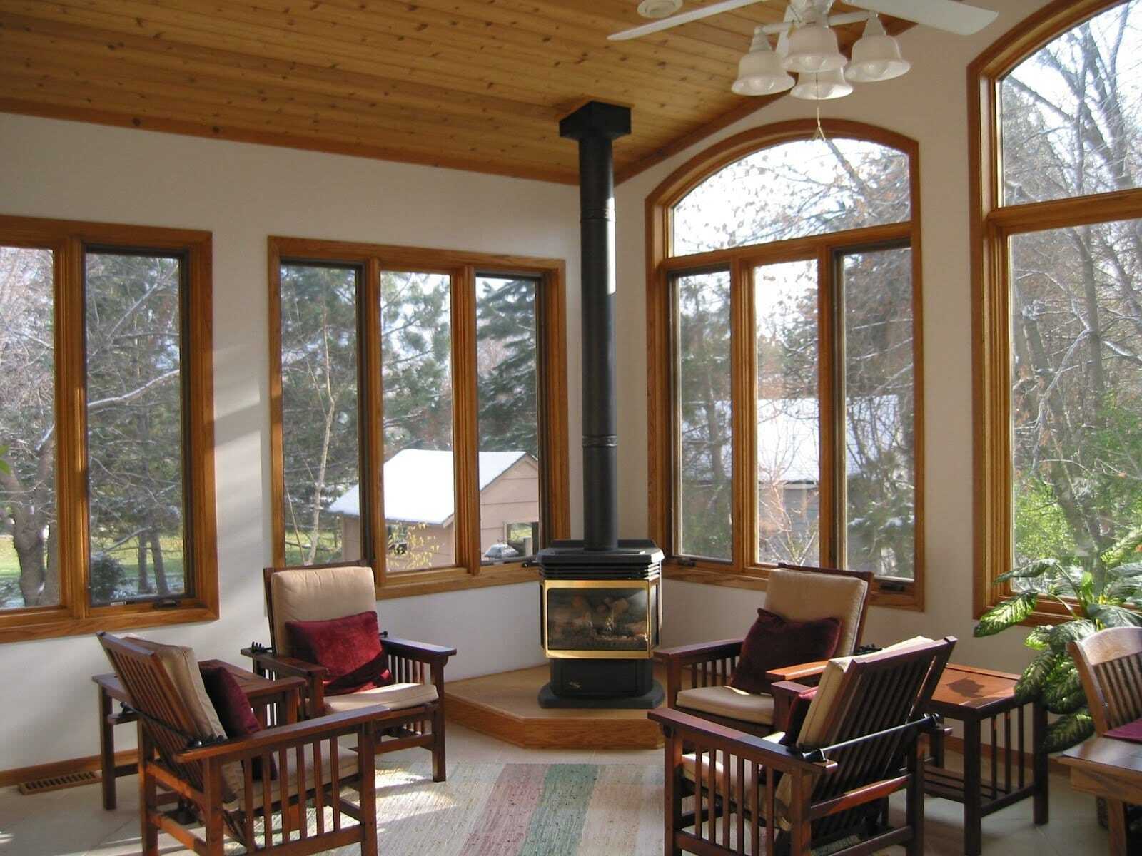 Просторная комната с окнами в деревянных рамах, деревянным потолком, камином и уютными креслами, вид на деревья за окном.