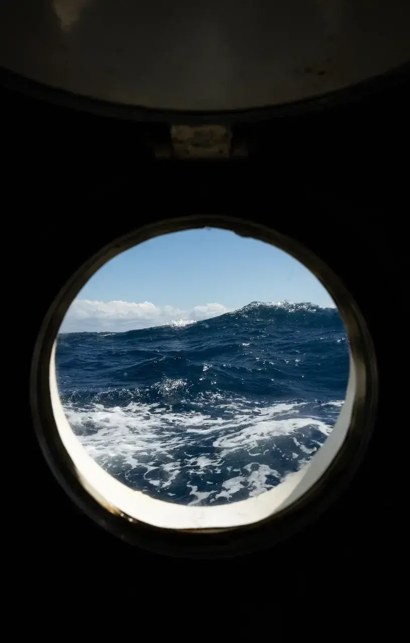 A view through the round porthole of a sailing yacht crossing the Drake Passage revealing powerful ocean swells and deep blue waves under a clear sky. The image captures the raw conditions of the Southern Ocean on the journey to Antarctica highlighting th