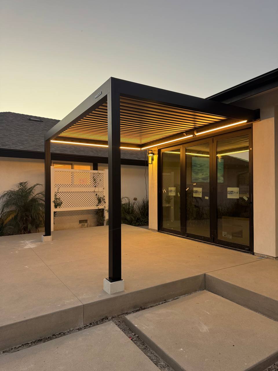 Warm roof lighting illuminates the patio beneath the black wall-mounted pergola.