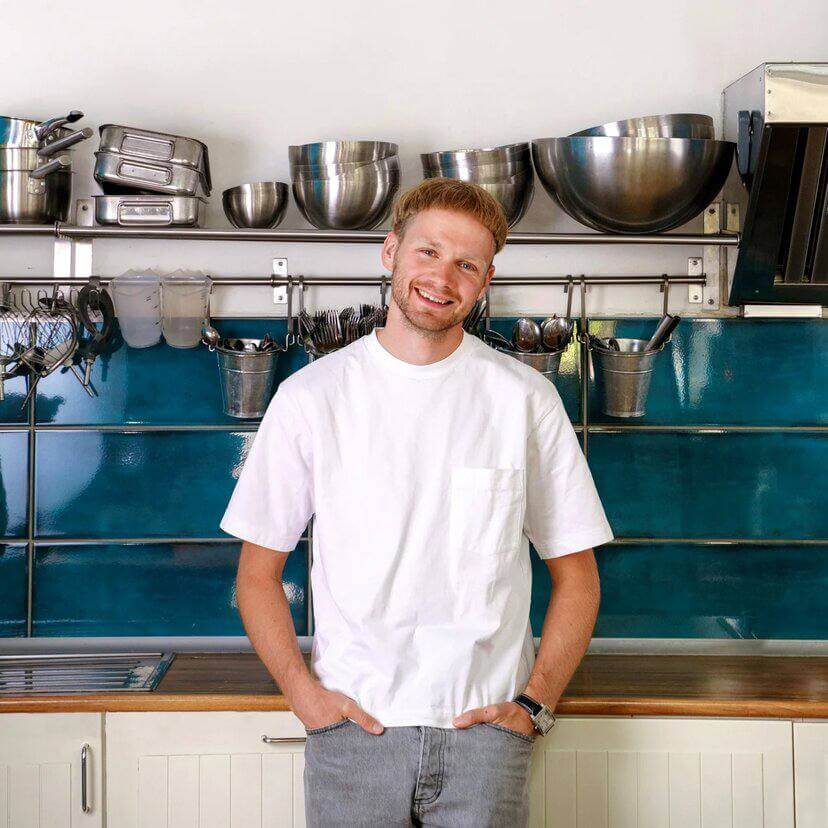 Портретная фотография улыбающегося мужчины со светлыми волосами, в белой футболке, стоящего на кухне с белыми деревянными шкафами, синей стеной и металлическими кухонными принадлежностями на заднем плане.