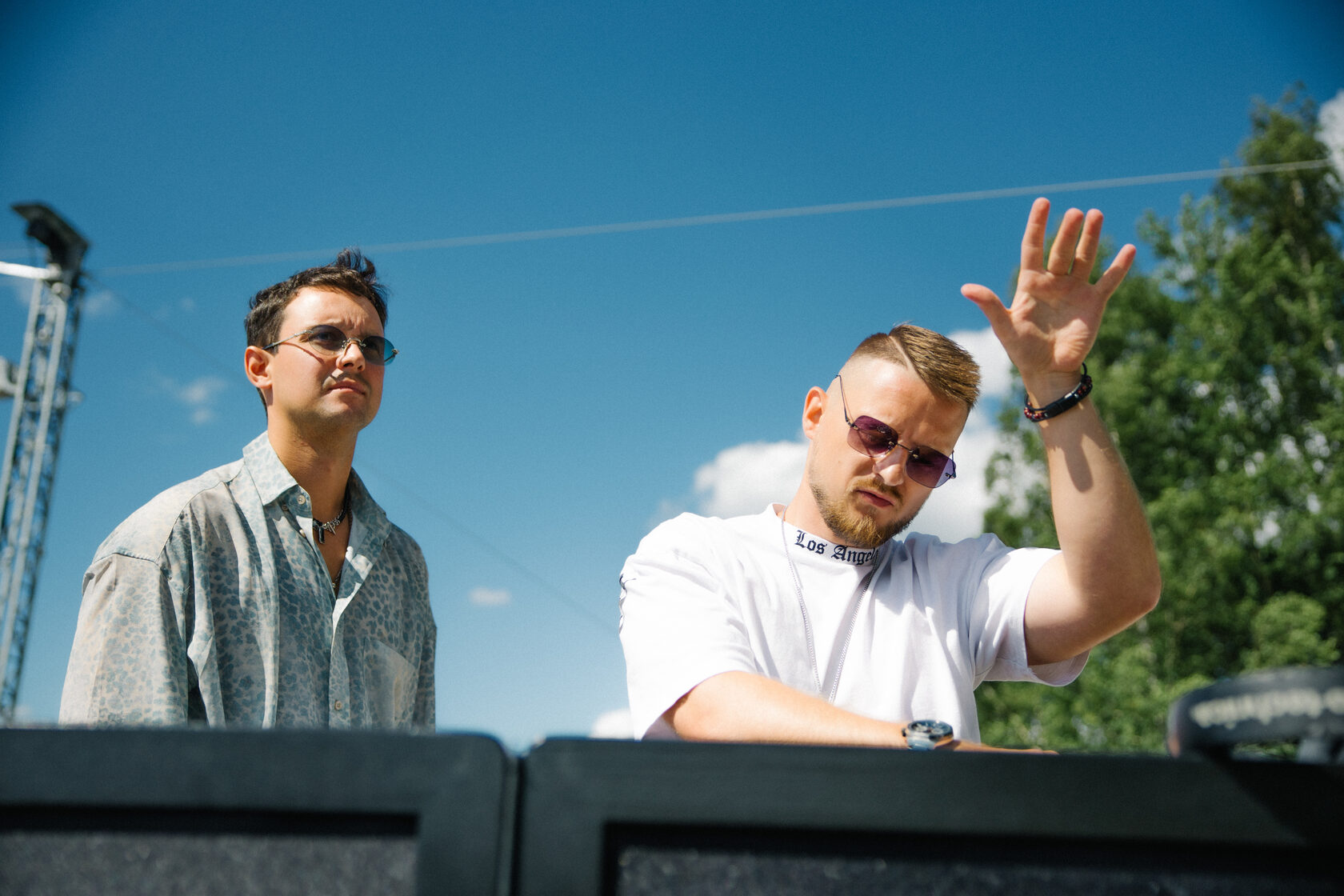 DJ ROZHKOV в белой рубашке поднимает руку за пультом на открытой площадке рядом с другим участником события.