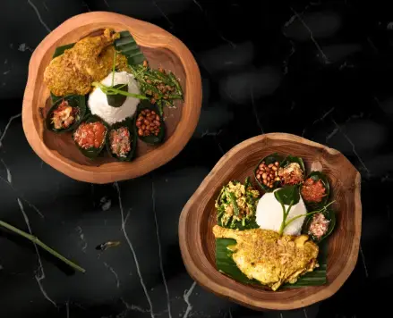 Two rustic wooden plates featuring poultry covered in a rich, traditional yellow Betutu spice blend. Served with white rice, green vegetables, and sambals, creating a uniquely Balinese Iftar in Ubud experience.