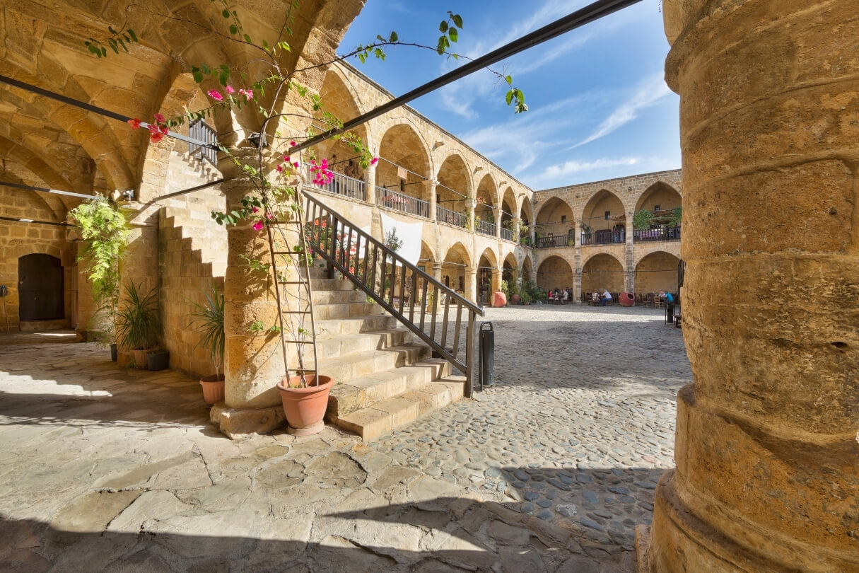 Внутренний двор старинного караван-сарая с арками и лестницей, украшенной цветами.
