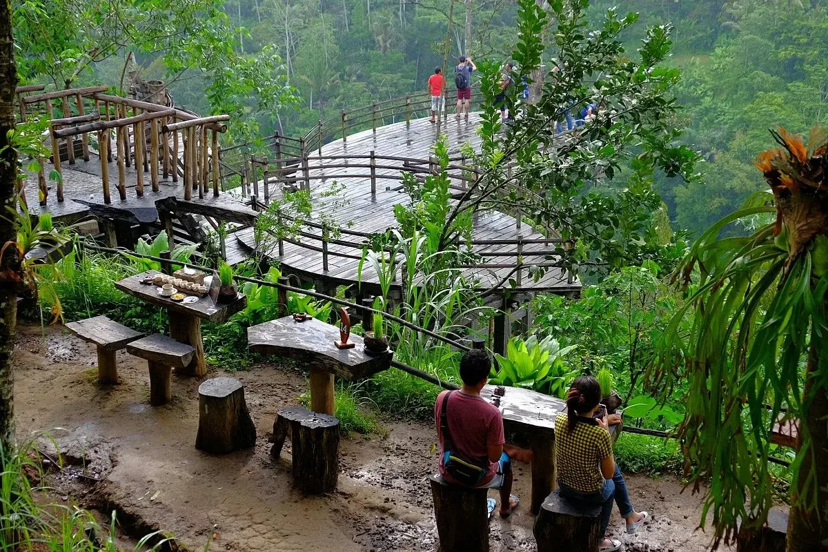 Visitors relaxing on a wooden viewing deck and outdoor cafe at Bali Pulina, enjoying authentic luwak coffee overlooking a lush green valley during an Ubud getaway.