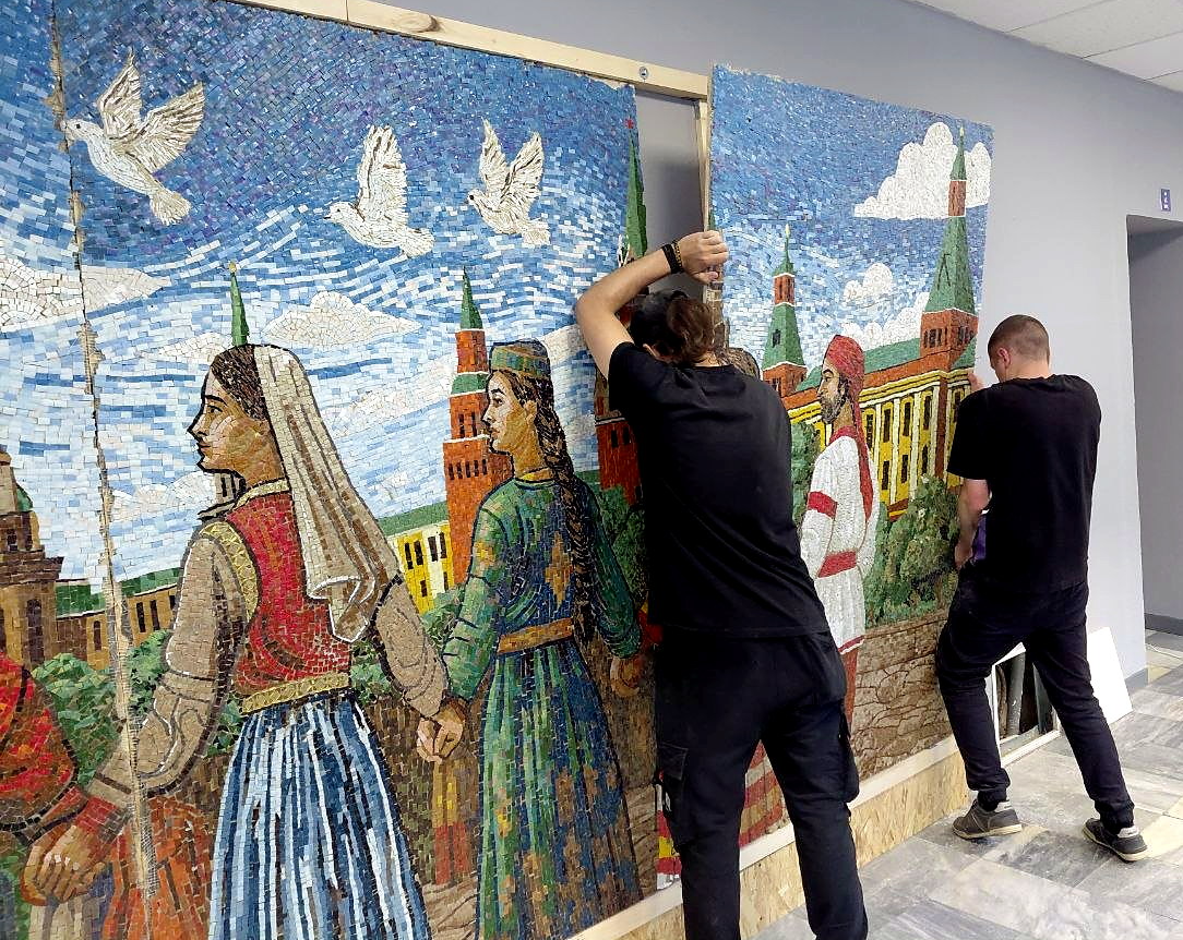 Двое мастеров монтируют фрагменты мозаики Дружба народов на стену с каркасом