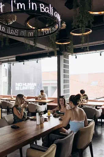 Friends gathering for food and drinks in the bright, cozy dining area of Murmur Lounge in Canggu, featuring a glowing neon Bali sign.