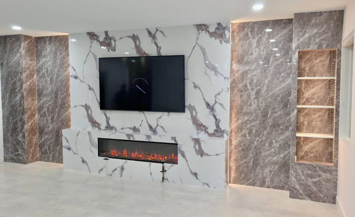 Completed modern media wall in Miami with marble-look panels, a mounted television, linear electric fireplace, LED lighting, and display shelving.