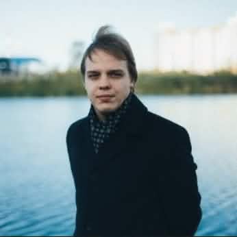 Портретная фотография молодого человека со светлыми волосами, с прической с легким начесом, в темном пальто и пестром шарфе, смотрящего влево, на фоне размытого водоема и зданий на заднем плане.