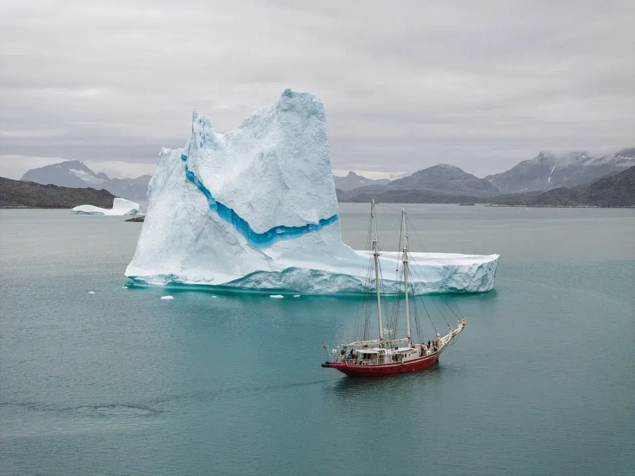 A classic expedition sailing yacht cruising beside a towering iceberg in the pristine fjords of East Greenland. The vivid blue ice, calm Arctic waters and rugged mountain backdrop highlight the scale of Greenland's glaciers and the unique experience of exploring the Arctic by small expedition yacht.