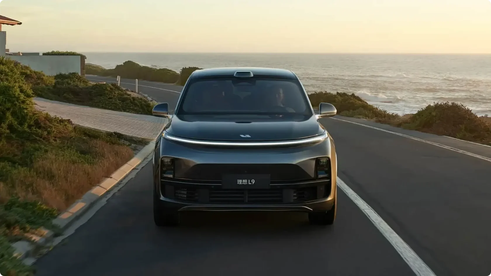 Движущийся по прибрежной дороге гибридный кроссовер Li Auto L9 текущего поколения в темно-сером цвете, фронтальный вид: светодиодная полоса по всей ширине, лидар на крыше и массивные воздухозаборники