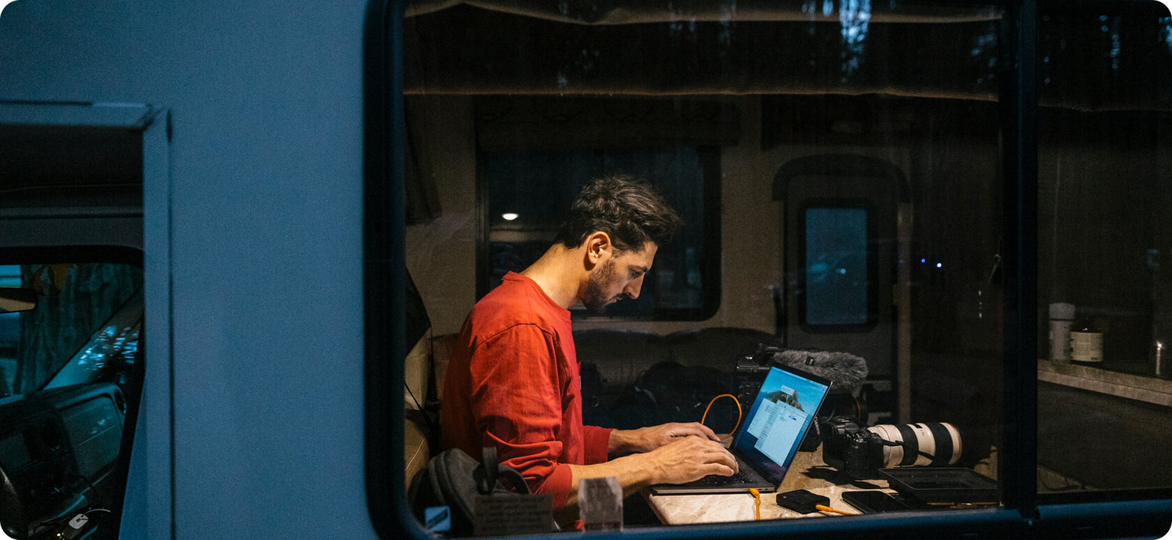 Persona escribiendo en un portátil sentada en la puerta abierta de una furgoneta camper con vistas a un valle montañoso.