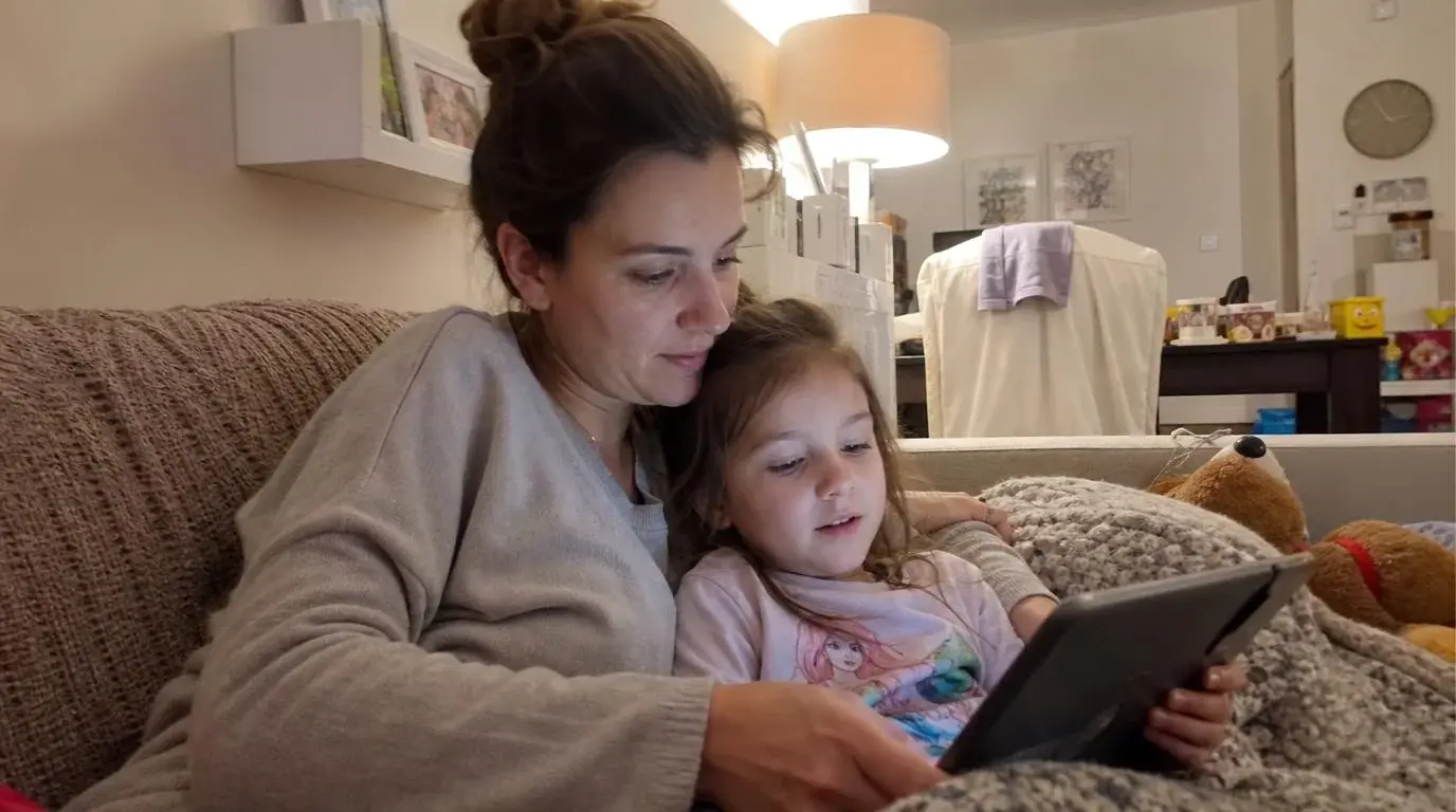 Mother hugging her toddler after healthy screen time, with an educational app playing on a tablet on the sofa, demonstrating offline skill transfer and no turn-off tantrums.