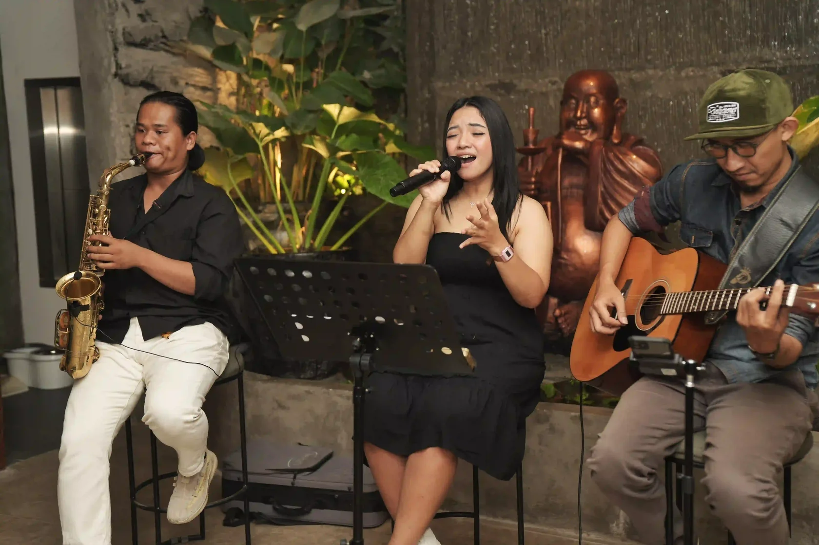 A talented trio of musicians consisting of a saxophonist, a female vocalist, and an acoustic guitarist performing live near a large bronze Buddha statue. For a relaxing coastal ambiance, this live music setting is arguably the best event in Bali.