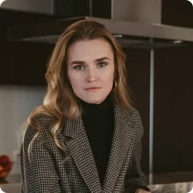 Портретная фотография молодой женщины со светлыми вьющимися волосами, с легким макияжем, в черной водолазке и сером клетчатом пиджаке, смотрящей прямо в камеру, на фоне темной кухни.