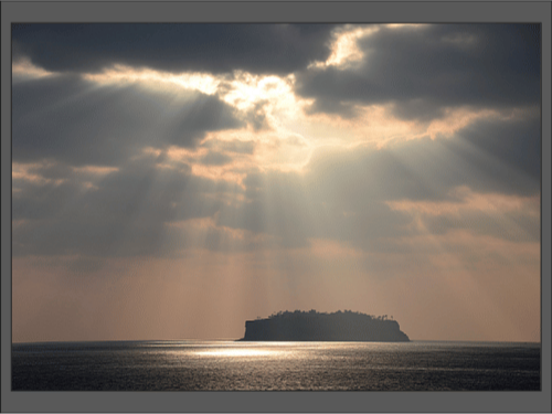 Солнечные лучи пробиваются сквозь облака и освещают маленький остров в море.