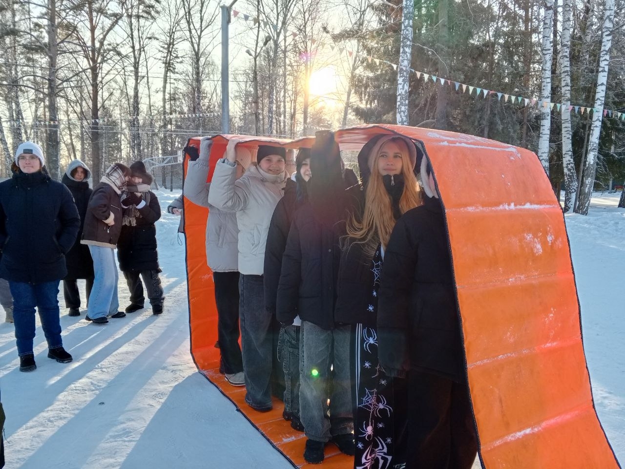 Дети в оранжевых пончо во время игры на территории центра, командный момент с берёзами и зимним пейзажем