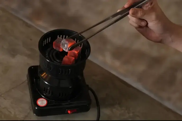 A close-up of a hand using metal tongs to carefully arrange glowing red coconut charcoal on an electric burner, the perfect preparation for a clean and smooth shisha smoke session.