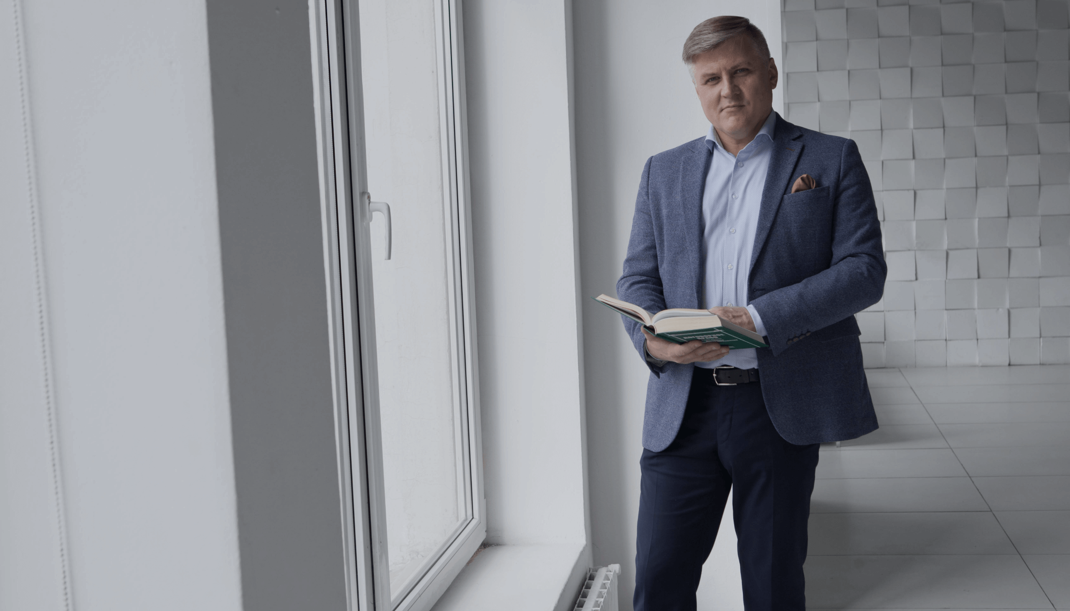 Мужчина в костюме стоит у окна с книгой в руках