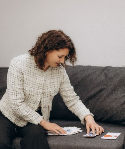 Женщина с кудрявыми волосами в светлом пиджаке сидит на диване и раскладывает бумаги или карточки на столе перед собой.