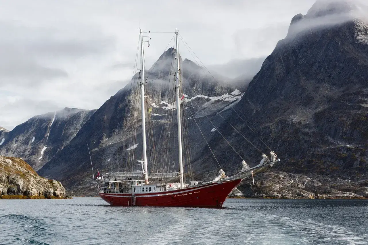 A classic expedition sailing yacht anchored in a remote East Greenland fjord surrounded by towering granite mountains dramatic cliffs and low Arctic clouds. The calm water reflects the rugged landscape showcasing the unique beauty of Greenlands untouched 