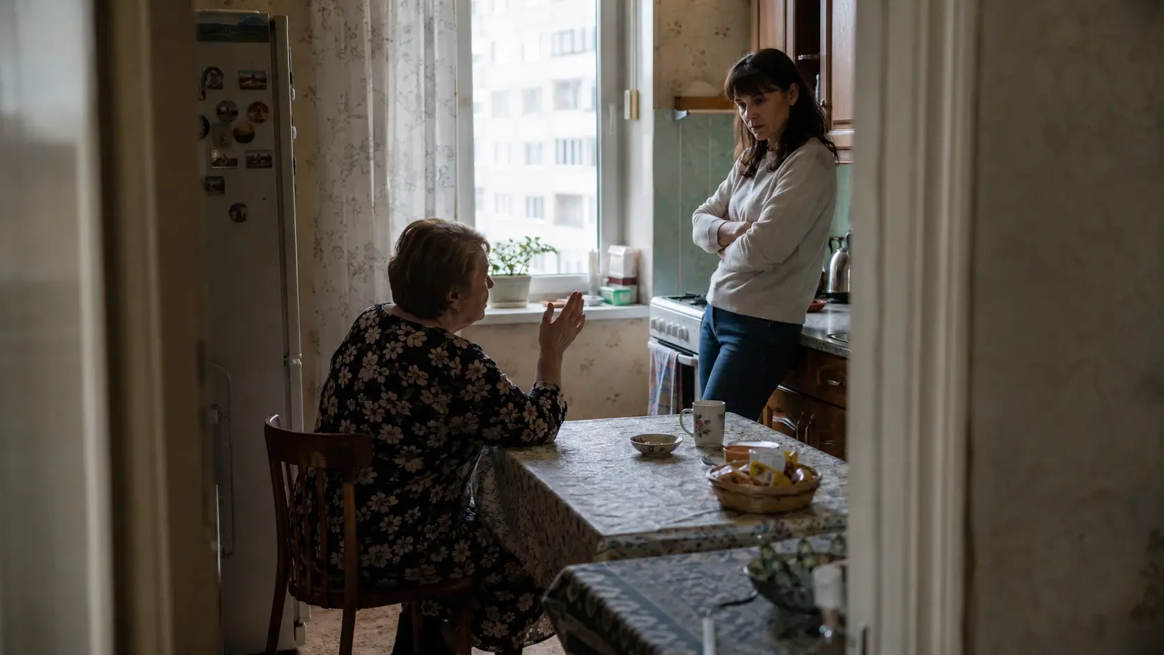 Женщина стоит у стола рядом с пожилой матерью, удерживая сложное чувство семейной вины.