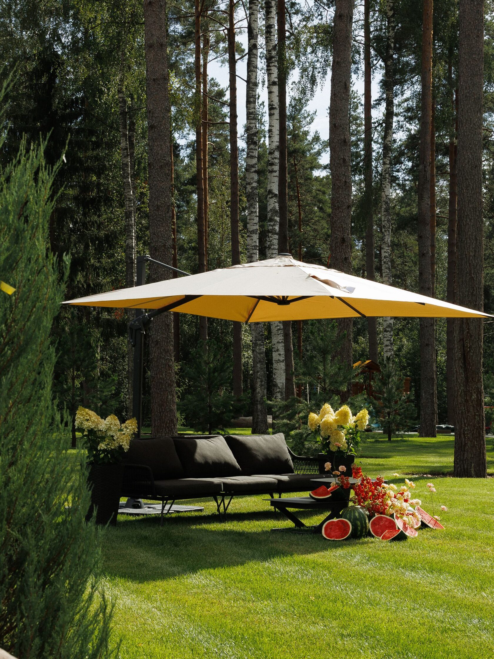 Загородная зона отдыха на территории клуба в сосновом лесу: чёрный диван под большим садовым зонтом, зелёный газон, цветочные композиции и арбуз на столике — атмосфера летней свадьбы или мероприятия за городом в СПб.