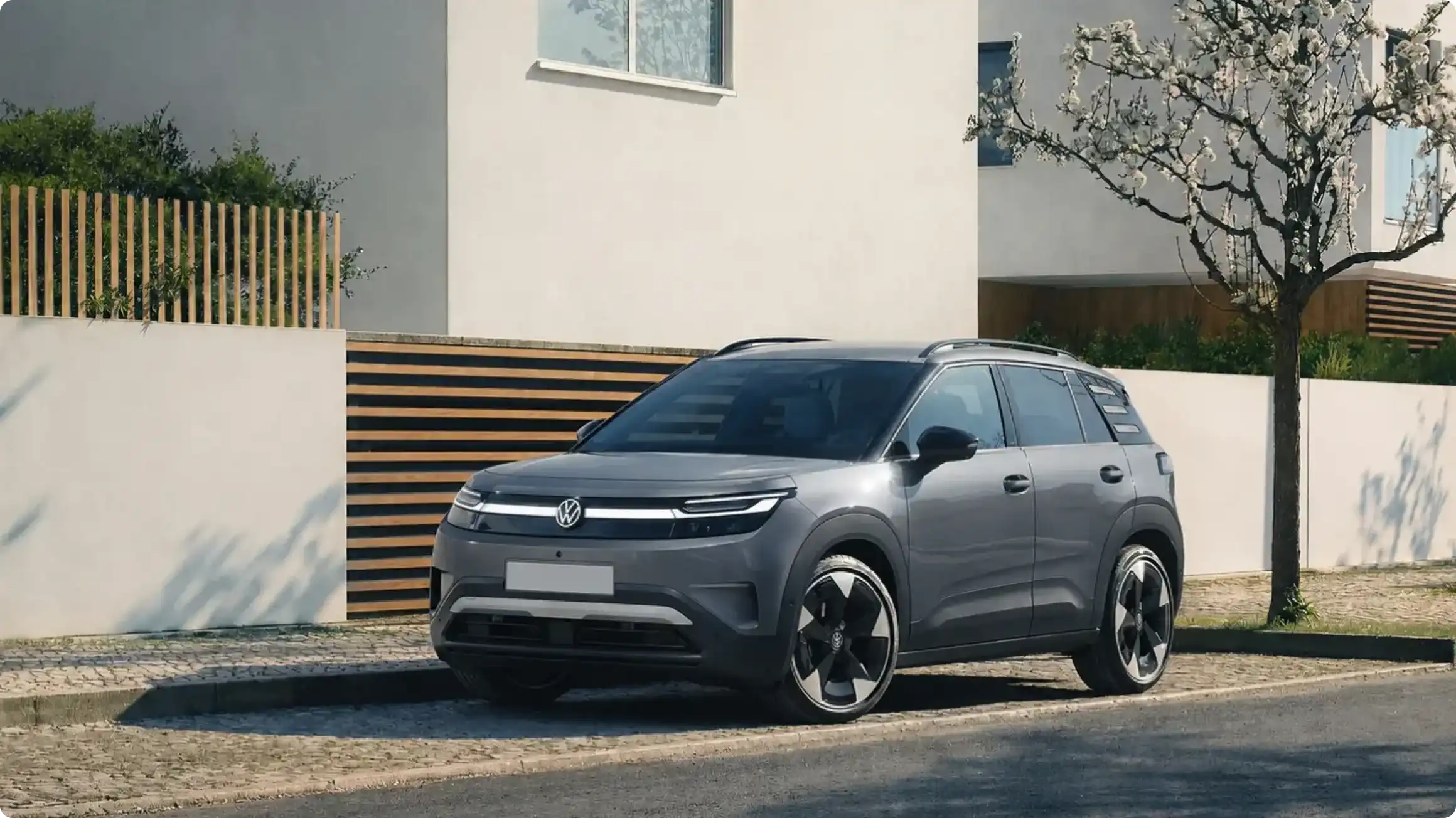 Volkswagen ID. Cross в сером цвете, вид спереди сбоку у жилого дома: светодиодная полоса с логотипом VW, черные молдинги и двухцветные диски.