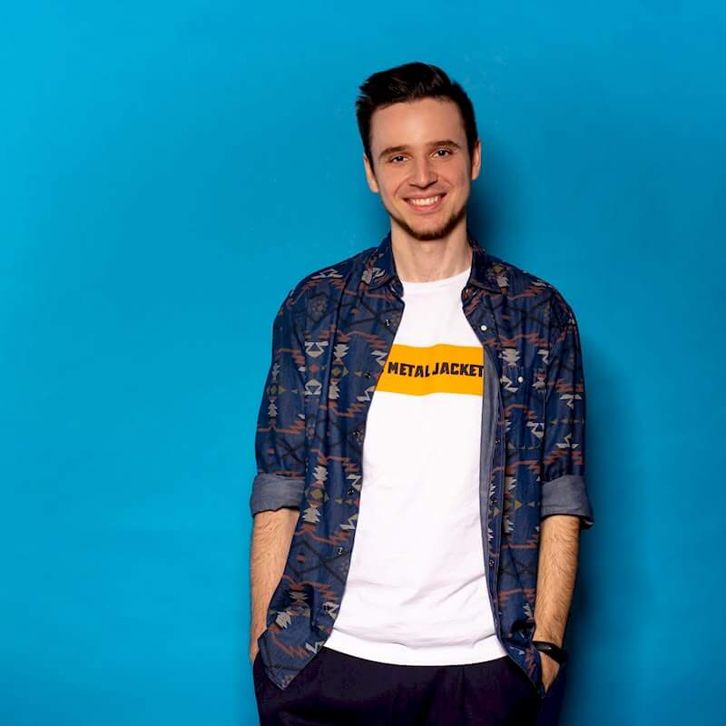 Портретная фотография улыбающегося молодого мужчины с темными волосами, с легкой небритостью, в синей рубашке с этническим узором поверх белой футболки с надписью 'METALJACKET', стоящего на фоне ярко-синей стены. Он смотрит прямо в камеру и руки держит в карманах.