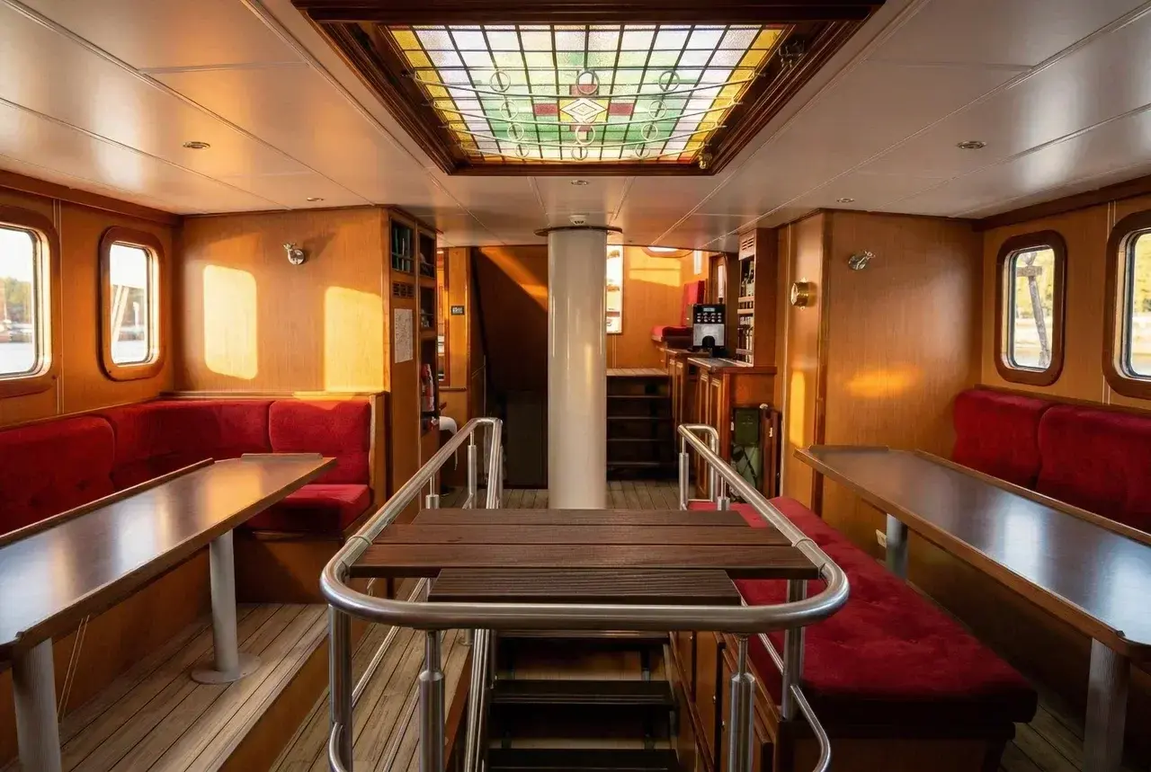 The spacious saloon aboard the schooner Eldorado, featuring warm wooden interiors, comfortable red seating areas, large dining tables, and a decorative stained-glass skylight. Sunlight fills the communal space, creating a cozy atmosphere where guests gather for meals, lectures, and conversations during polar expeditions and ocean crossings.