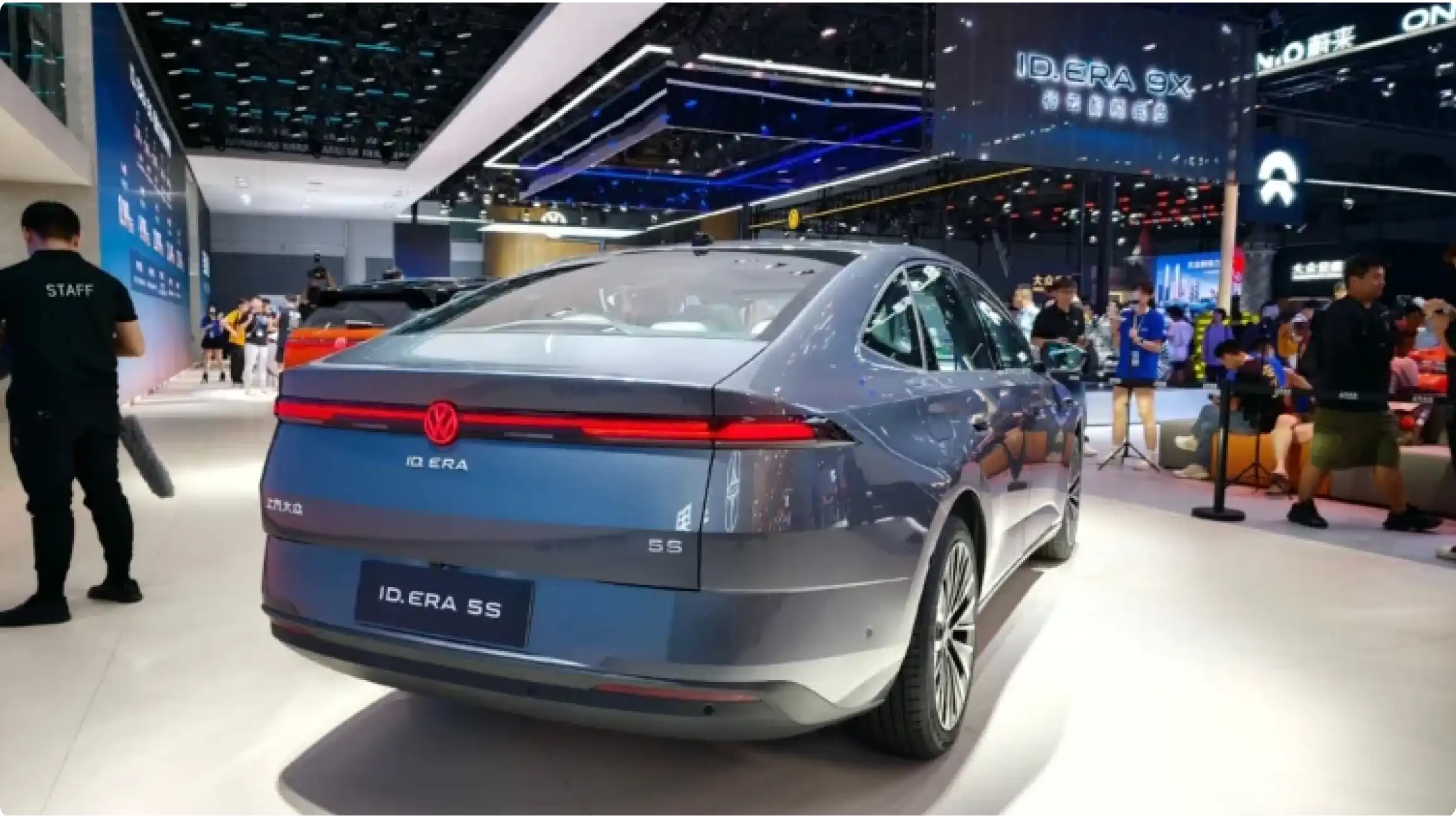 Задняя часть гибридного седана Volkswagen ID. Era 5S в синем цвете на автосалоне в Чэнду: светодиодная полоса, номерной знак ID. ERA 5S и маркировка 5S на багажнике.
