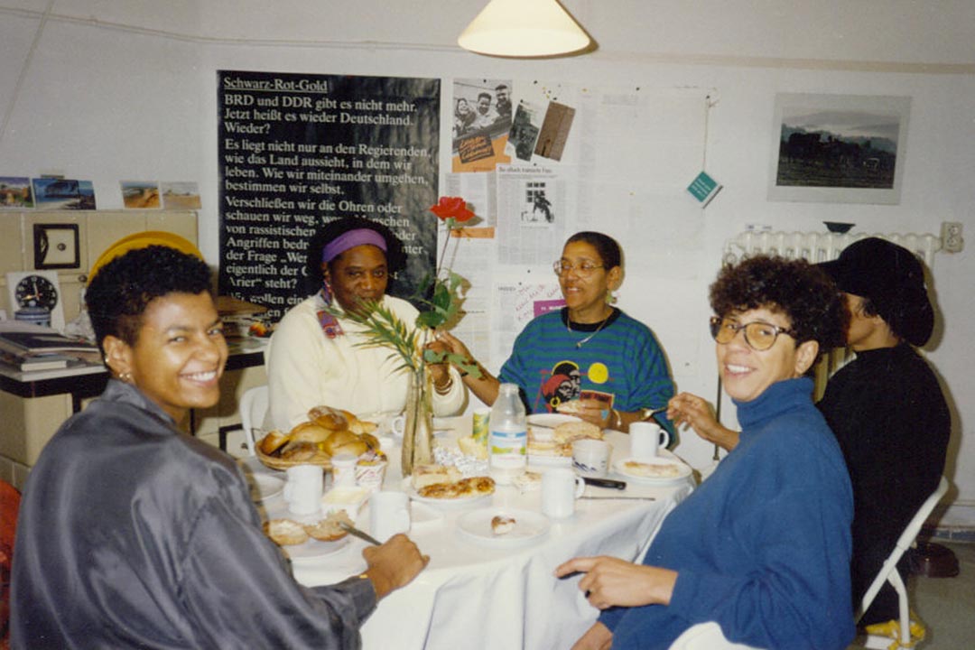 Тифлокомментарий. Цветная фотография. За круглым столом пять женщин пьют чай. Это Одри Лорд и сотрудницы издательства «Орланда». Стол покрыт белой скатертью и уставлен чашками и тарелками с выпечкой. В центре стола красная роза в узкой вазе. Одри обернулась и широко улыбается. В комнате на белых стенах развешены плакаты, фотографии и листовки. 