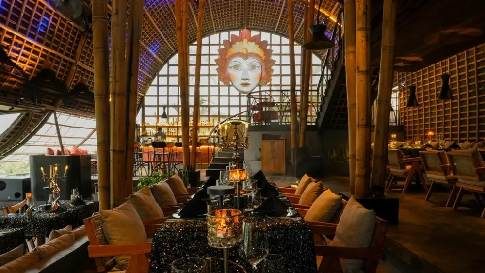 An unforgettable top dinner in Ubud inside an impressive organic bamboo structure, featuring elegant dark table settings, warm candlelight, and a magnificent Balinese mask decoration on the wall.