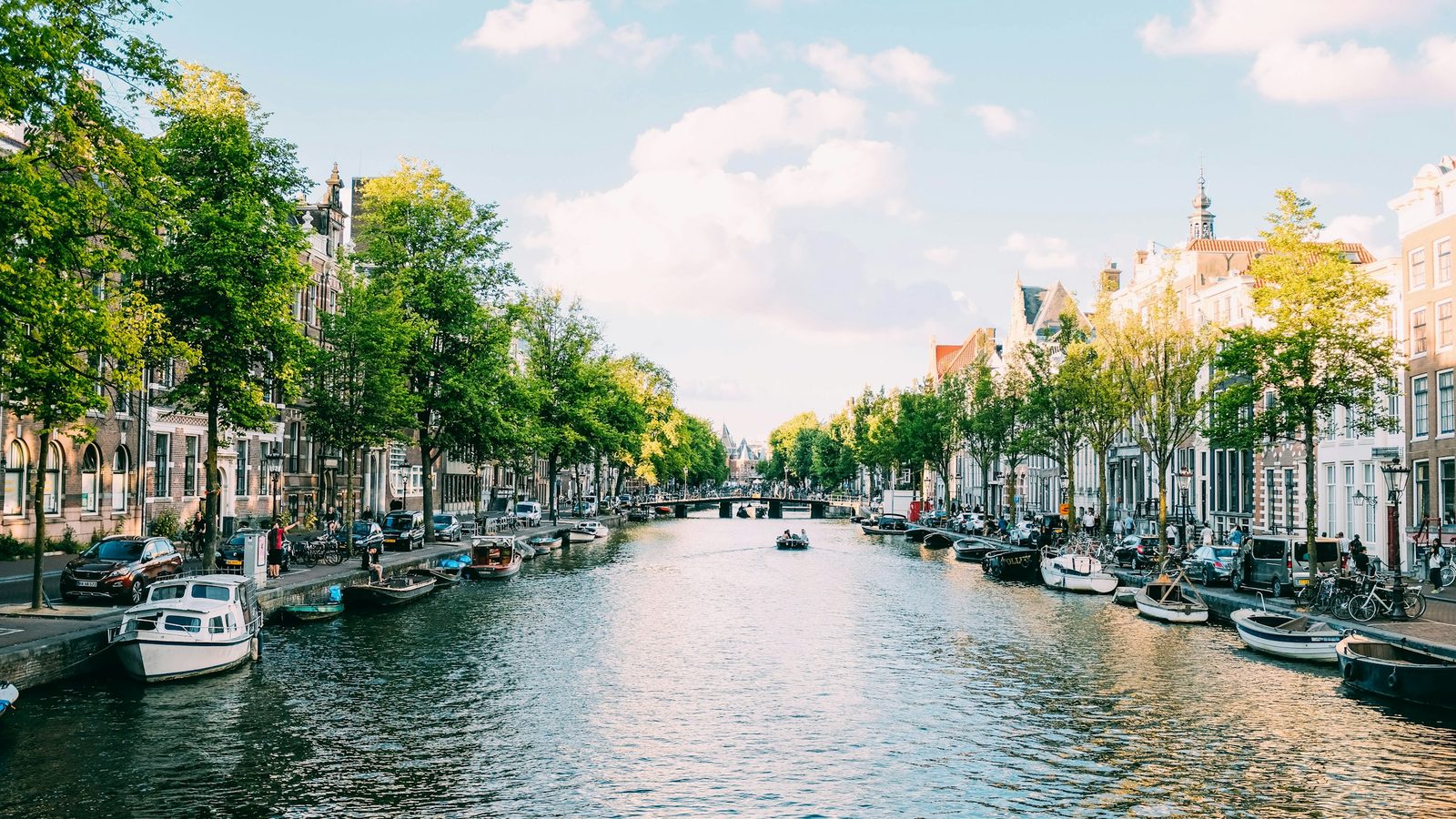 Boats moored along a tree-lined Amsterdam canal with historic canal houses on both sides and a bridge in the distance