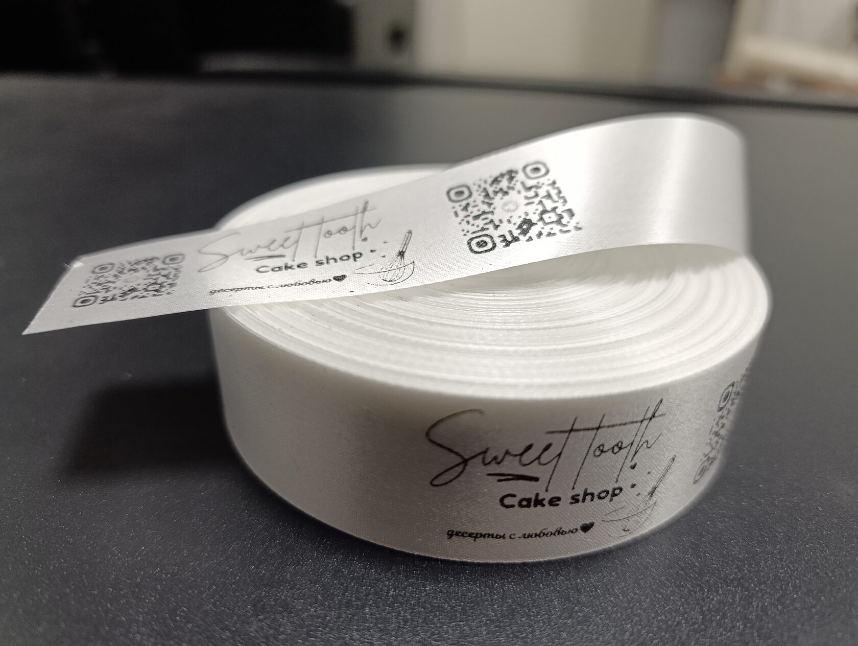 На изображении рулон белой ленты с надписью «Sweet Note Cake shop». Сверху лежит этикетка с QR-кодом и логотипом.