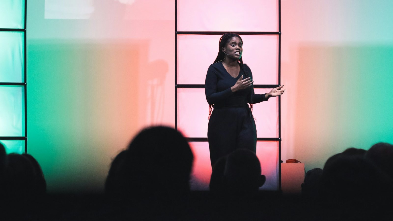 Woman speaking on a stage with a headset microphone in front of a pink and green lit backdrop, audience in silhouette