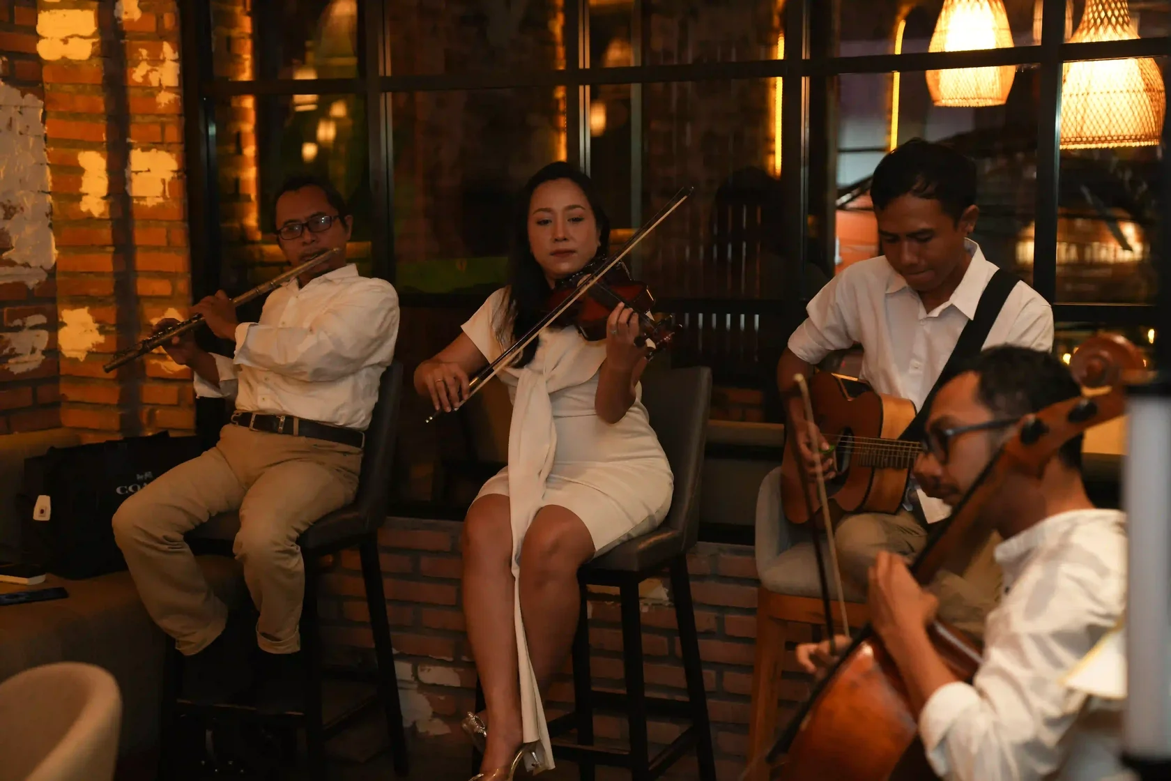 A refined live music quartet featuring a flute, violin, acoustic guitar, and cello playing against a warm brick wall backdrop. Experiencing sophisticated live tunes like this is often considered the best event in Bali for music lovers.