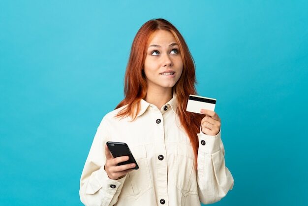 Teenager Russian girl isolated on blue wall buying with the mobile with a credit card while thinking