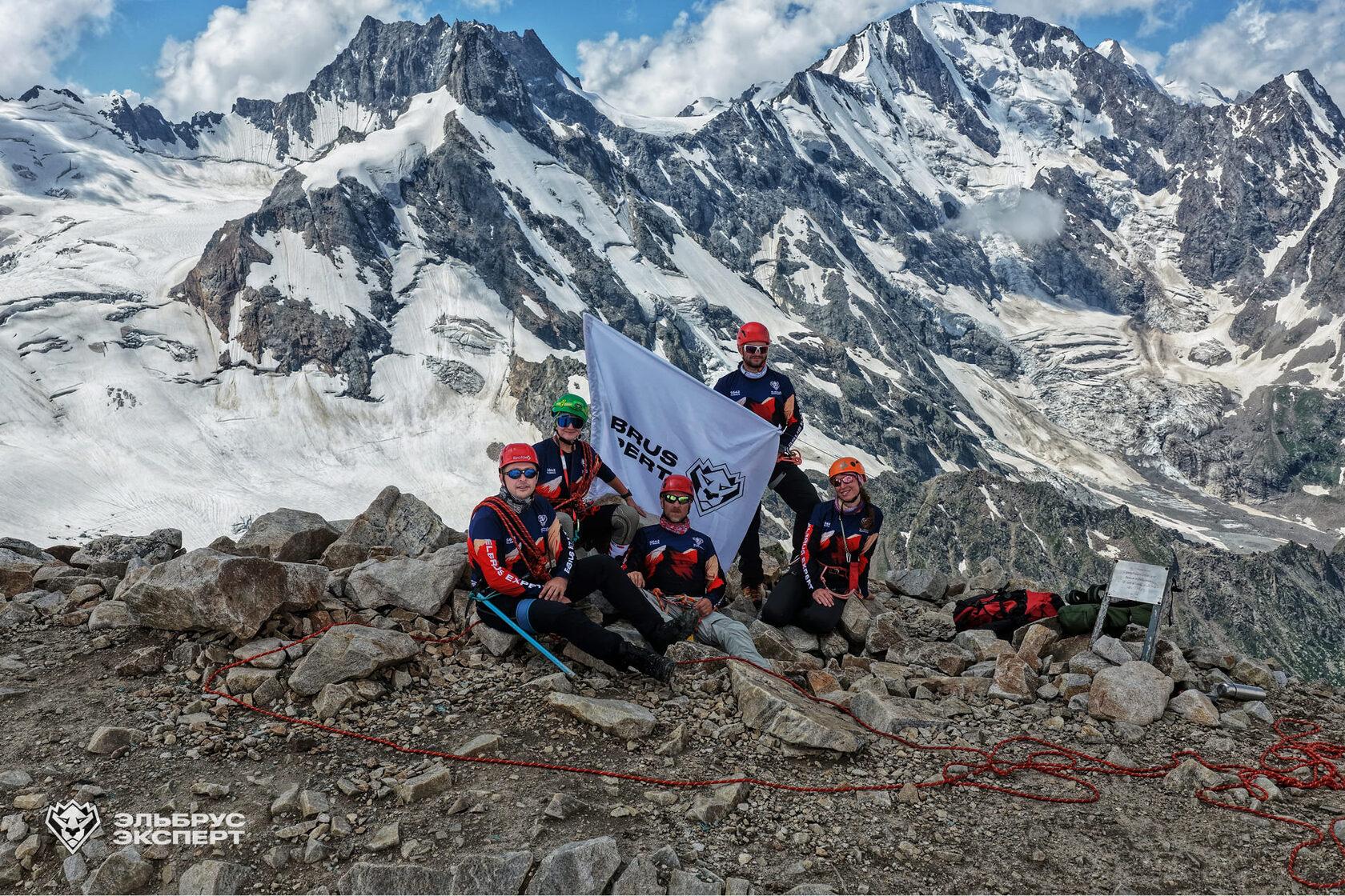 Course group with an Elbrus Expert flag on a rocky ridge in front of a glacier-covered mountain range
