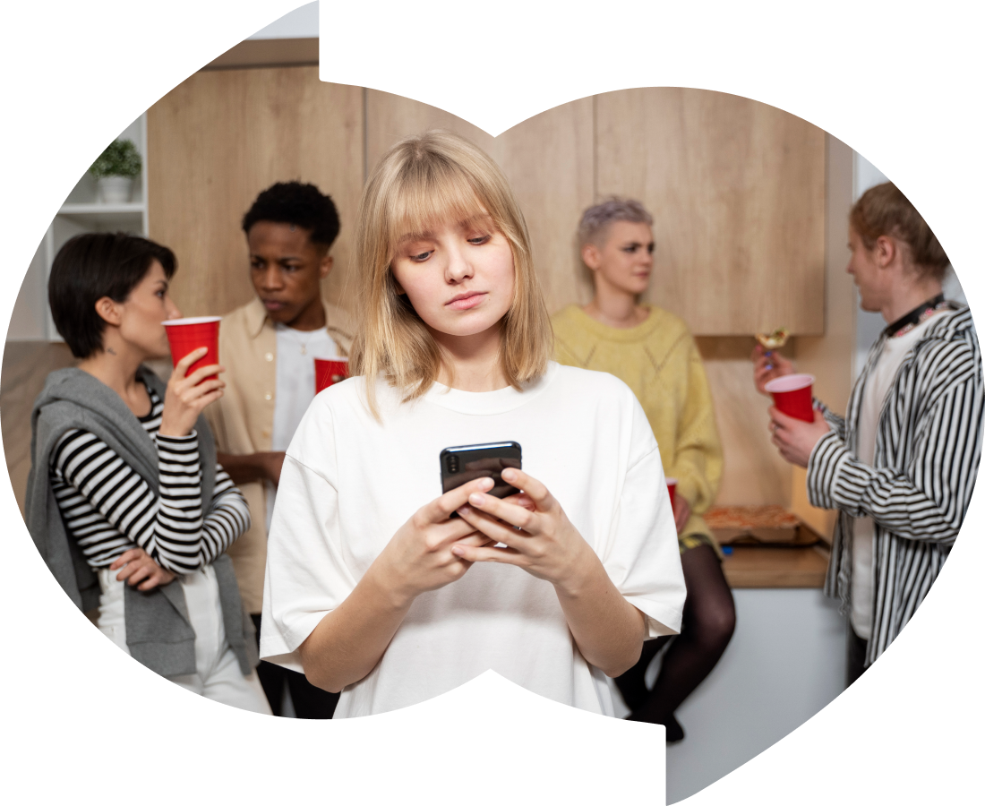 A young woman stands in the foreground, focused on her phone, while a group of four people socialize in the background. They are gathered in a kitchen, holding red cups and chatting. The scene suggests a social gathering, but the woman appears detached or